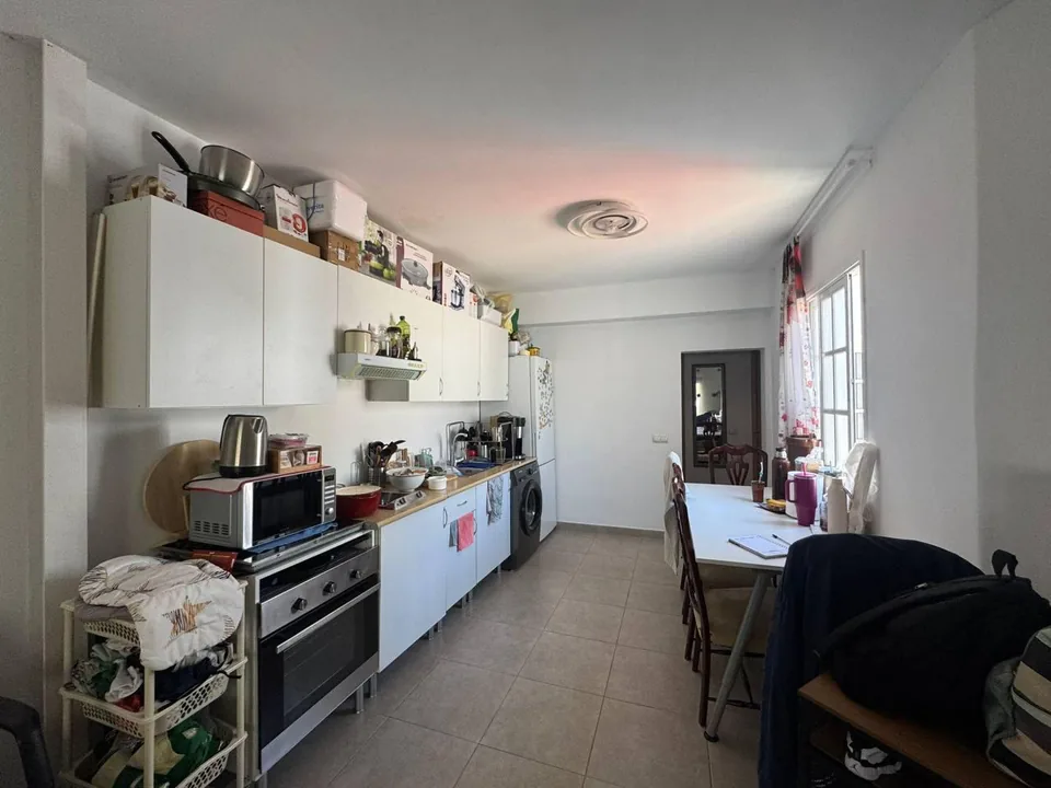 Cocina antes de la reforma, con mobiliario blanco, electrodomésticos y mesa junto a la ventana